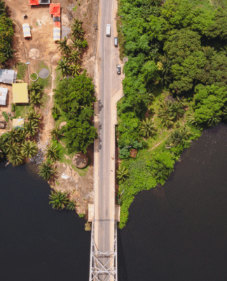 Imagem aérea mostra uma ponte estreita cruzando um rio escuro, ligando duas margens contrastantes — à esquerda, uma área urbana com casas simples e telhados coloridos; à direita, uma região de vegetação densa e verde. A cena evidencia a transição entre ambiente urbano e natureza.