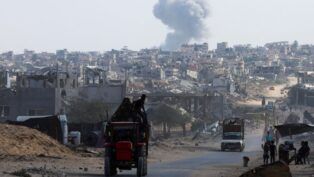 Trator carrega duas pessoas em estrada enquanto ao fundo uma fumaça grande aparece entre as casas