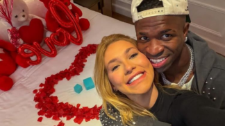 Vinícius Júnior e Virginia estão abraçados à frente de uma cama com pétalas de rosa e balões vermelhos