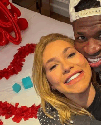 Vinícius Júnior e Virginia estão abraçados à frente de uma cama com pétalas de rosa e balões vermelhos