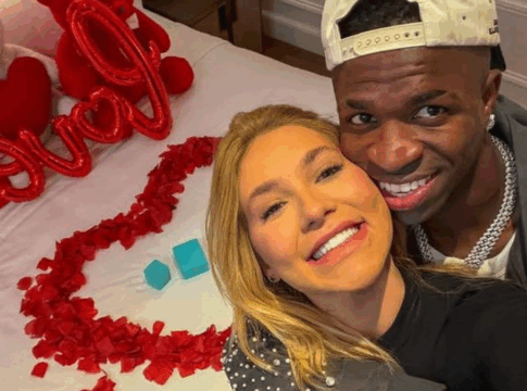 Vinícius Júnior e Virginia estão abraçados à frente de uma cama com pétalas de rosa e balões vermelhos