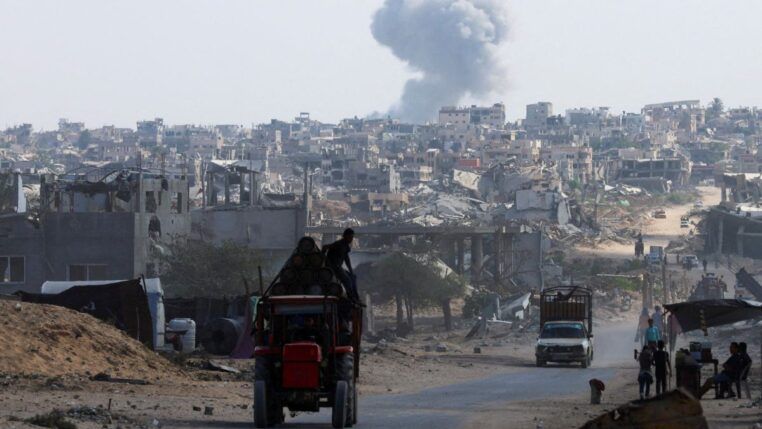 Trator carrega duas pessoas em estrada enquanto ao fundo uma fumaça grande aparece entre as casas