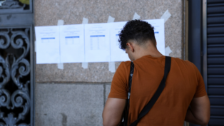 Homem de camiseta laranja e bolsa transversal preta observa folhas impressas afixadas com fita adesiva em uma parede de pedra. Os papéis contêm listas com nomes e números, possivelmente resultados ou locais de prova. A cena ocorre em ambiente urbano, em frente a um prédio público, com parte de um portão de ferro decorado visível à esquerda.