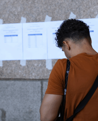 Homem de camiseta laranja e bolsa transversal preta observa folhas impressas afixadas com fita adesiva em uma parede de pedra. Os papéis contêm listas com nomes e números, possivelmente resultados ou locais de prova. A cena ocorre em ambiente urbano, em frente a um prédio público, com parte de um portão de ferro decorado visível à esquerda.