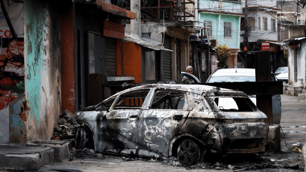 Homem está atrás de carro destruído por fogo na entrada da Favela da Penha