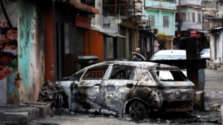 Homem está atrás de carro destruído por fogo na entrada da Favela da Penha