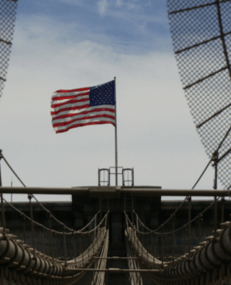 Fotografia tirada de uma ponte suspensa com cabos de aço e grades metálicas convergindo para o centro da imagem. Ao fundo, no ponto mais alto da estrutura, tremula uma bandeira dos Estados Unidos sob um céu parcialmente nublado. A composição destaca as linhas simétricas da ponte e o contraste entre o aço e o movimento suave da bandeira.