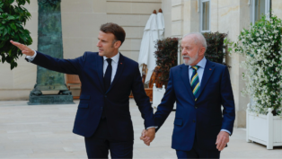 Dois homens de terno escuro estão de mãos dadas e caminhando em uma área externa elegante, com plantas e paredes de pedra clara ao fundo. O homem à esquerda gesticula com o braço estendido, apontando algo à frente, enquanto o homem à direita olha na mesma direção com expressão atenta. Ambos parecem estar em um ambiente formal e amistoso.