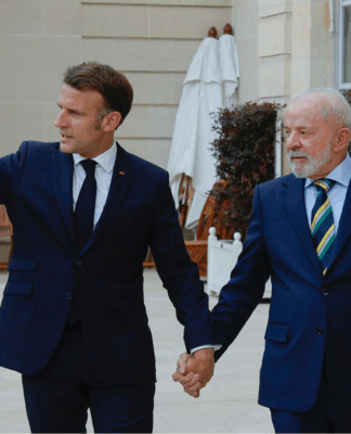 Dois homens de terno escuro estão de mãos dadas e caminhando em uma área externa elegante, com plantas e paredes de pedra clara ao fundo. O homem à esquerda gesticula com o braço estendido, apontando algo à frente, enquanto o homem à direita olha na mesma direção com expressão atenta. Ambos parecem estar em um ambiente formal e amistoso.