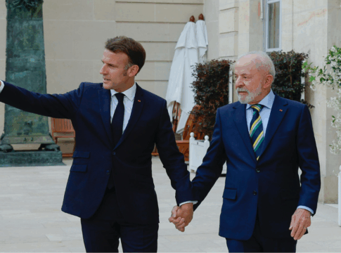 Dois homens de terno escuro estão de mãos dadas e caminhando em uma área externa elegante, com plantas e paredes de pedra clara ao fundo. O homem à esquerda gesticula com o braço estendido, apontando algo à frente, enquanto o homem à direita olha na mesma direção com expressão atenta. Ambos parecem estar em um ambiente formal e amistoso.