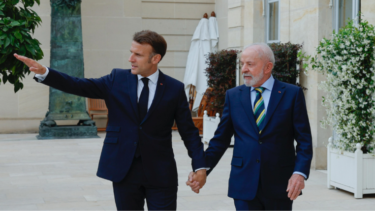 Dois homens de terno escuro estão de mãos dadas e caminhando em uma área externa elegante, com plantas e paredes de pedra clara ao fundo. O homem à esquerda gesticula com o braço estendido, apontando algo à frente, enquanto o homem à direita olha na mesma direção com expressão atenta. Ambos parecem estar em um ambiente formal e amistoso.