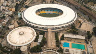 Imagem aérea de um grande complexo esportivo urbano. No centro, destaca-se um estádio de futebol de formato oval, com cobertura branca e arquibancadas coloridas em tons de amarelo e azul. À esquerda, há uma estrutura circular coberta, semelhante a uma arena. À direita, vê-se uma área com duas piscinas retangulares, uma delas olímpica. O entorno é composto por ruas, árvores e prédios residenciais, além de linhas de trem ao fundo.