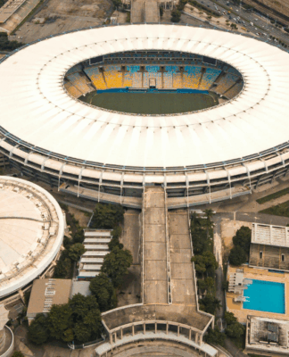 Imagem aérea de um grande complexo esportivo urbano. No centro, destaca-se um estádio de futebol de formato oval, com cobertura branca e arquibancadas coloridas em tons de amarelo e azul. À esquerda, há uma estrutura circular coberta, semelhante a uma arena. À direita, vê-se uma área com duas piscinas retangulares, uma delas olímpica. O entorno é composto por ruas, árvores e prédios residenciais, além de linhas de trem ao fundo.
