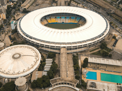 Imagem aérea de um grande complexo esportivo urbano. No centro, destaca-se um estádio de futebol de formato oval, com cobertura branca e arquibancadas coloridas em tons de amarelo e azul. À esquerda, há uma estrutura circular coberta, semelhante a uma arena. À direita, vê-se uma área com duas piscinas retangulares, uma delas olímpica. O entorno é composto por ruas, árvores e prédios residenciais, além de linhas de trem ao fundo.