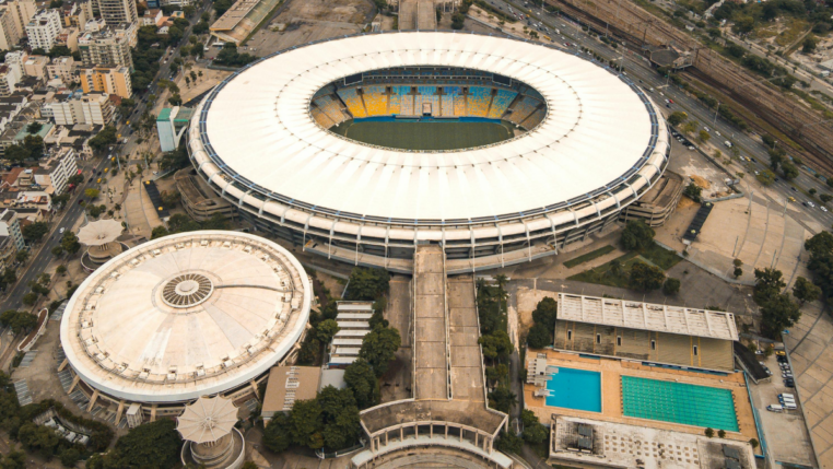 Imagem aérea de um grande complexo esportivo urbano. No centro, destaca-se um estádio de futebol de formato oval, com cobertura branca e arquibancadas coloridas em tons de amarelo e azul. À esquerda, há uma estrutura circular coberta, semelhante a uma arena. À direita, vê-se uma área com duas piscinas retangulares, uma delas olímpica. O entorno é composto por ruas, árvores e prédios residenciais, além de linhas de trem ao fundo.