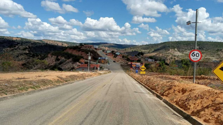 Radar de controle de velocidade instalado em rodovia de área rural, próximo a vegetação e montes, em um dia ensolarado.