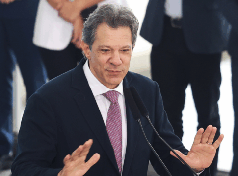 Homem de terno azul escuro, camisa branca e gravata rosa fala em um púlpito com dois microfones. Ele gesticula com as mãos levantadas à altura do peito, como se enfatizasse um ponto. Ao fundo, pessoas vestidas formalmente aparecem desfocadas, sugerindo um ambiente oficial ou de evento público.