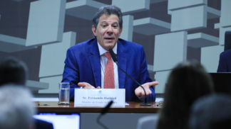 Homem de terno azul e gravata vermelha fala ao microfone em uma mesa de audiência. À sua frente há uma placa com o nome “Sr. Fernando Haddad, Ministro de Estado da Fazenda”. Ele gesticula com as mãos abertas, em gesto de explicação. Ao fundo, há um painel com formas geométricas e pessoas assistindo à fala.