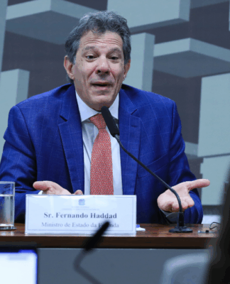 Homem de terno azul e gravata vermelha fala ao microfone em uma mesa de audiência. À sua frente há uma placa com o nome “Sr. Fernando Haddad, Ministro de Estado da Fazenda”. Ele gesticula com as mãos abertas, em gesto de explicação. Ao fundo, há um painel com formas geométricas e pessoas assistindo à fala.