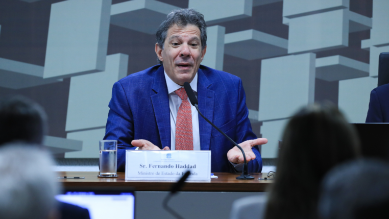 Homem de terno azul e gravata vermelha fala ao microfone em uma mesa de audiência. À sua frente há uma placa com o nome “Sr. Fernando Haddad, Ministro de Estado da Fazenda”. Ele gesticula com as mãos abertas, em gesto de explicação. Ao fundo, há um painel com formas geométricas e pessoas assistindo à fala.