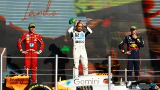 Três pilotos de Fórmula 1 estão no pódio após uma corrida. No centro, o vencedor, vestido com macacão branco da equipe McLaren, segura o boné verde da Pirelli sobre a cabeça. À esquerda, um piloto da Ferrari, de macacão vermelho, observa com as mãos na cintura. À direita, um piloto da Red Bull, de macacão azul escuro, mantém a mão no queixo. Em primeiro plano, parte do carro laranja e preto da McLaren aparece, e ao fundo, um telão exibe o nome “Lando”, indicando o vencedor da prova.