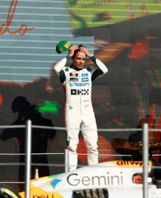 Três pilotos de Fórmula 1 estão no pódio após uma corrida. No centro, o vencedor, vestido com macacão branco da equipe McLaren, segura o boné verde da Pirelli sobre a cabeça. À esquerda, um piloto da Ferrari, de macacão vermelho, observa com as mãos na cintura. À direita, um piloto da Red Bull, de macacão azul escuro, mantém a mão no queixo. Em primeiro plano, parte do carro laranja e preto da McLaren aparece, e ao fundo, um telão exibe o nome “Lando”, indicando o vencedor da prova.