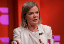 Mulher de cabelos loiros curtos veste blazer branco e fala durante uma entrevista ou debate em estúdio com fundo vermelho e luzes roxas. Ela gesticula com as mãos e usa microfone preso à roupa.