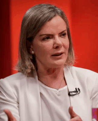 Mulher de cabelos loiros curtos veste blazer branco e fala durante uma entrevista ou debate em estúdio com fundo vermelho e luzes roxas. Ela gesticula com as mãos e usa microfone preso à roupa.