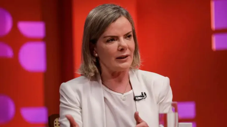 Mulher de cabelos loiros curtos veste blazer branco e fala durante uma entrevista ou debate em estúdio com fundo vermelho e luzes roxas. Ela gesticula com as mãos e usa microfone preso à roupa.