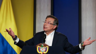 Homem de terno escuro e camisa branca fala em um púlpito com o brasão da Colômbia. Ele está com os braços abertos e expressão séria, olhando para o alto. Usa óculos e várias pulseiras coloridas nos pulsos. Ao fundo, há um painel com as cores da bandeira colombiana — amarelo, azul e vermelho —, sugerindo um pronunciamento oficial.