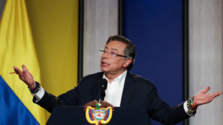 Homem de terno escuro e camisa branca fala em um púlpito com o brasão da Colômbia. Ele está com os braços abertos e expressão séria, olhando para o alto. Usa óculos e várias pulseiras coloridas nos pulsos. Ao fundo, há um painel com as cores da bandeira colombiana — amarelo, azul e vermelho —, sugerindo um pronunciamento oficial.