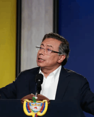 Homem de terno escuro e camisa branca fala em um púlpito com o brasão da Colômbia. Ele está com os braços abertos e expressão séria, olhando para o alto. Usa óculos e várias pulseiras coloridas nos pulsos. Ao fundo, há um painel com as cores da bandeira colombiana — amarelo, azul e vermelho —, sugerindo um pronunciamento oficial.