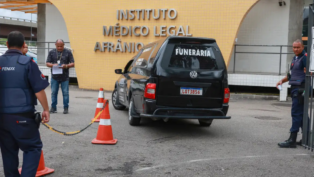 Carro funerário preto com a palavra “FUNERÁRIA” estaciona em frente ao prédio do Instituto Médico Legal Afrânio Peixoto, no Rio de Janeiro. Três homens, incluindo um policial e um agente de segurança, estão próximos ao veículo, enquanto cones laranja delimitam a área de acesso