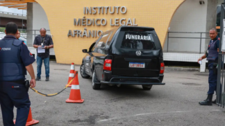 Carro funerário preto com a palavra “FUNERÁRIA” estaciona em frente ao prédio do Instituto Médico Legal Afrânio Peixoto, no Rio de Janeiro. Três homens, incluindo um policial e um agente de segurança, estão próximos ao veículo, enquanto cones laranja delimitam a área de acesso