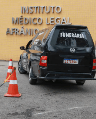 Carro funerário preto com a palavra “FUNERÁRIA” estaciona em frente ao prédio do Instituto Médico Legal Afrânio Peixoto, no Rio de Janeiro. Três homens, incluindo um policial e um agente de segurança, estão próximos ao veículo, enquanto cones laranja delimitam a área de acesso