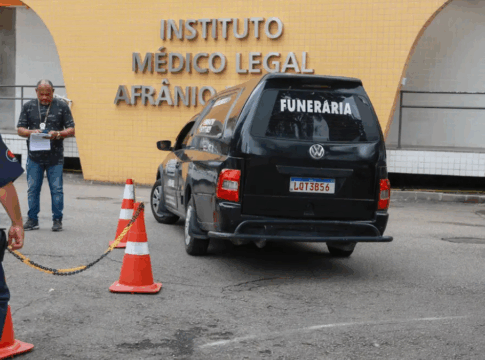 Carro funerário preto com a palavra “FUNERÁRIA” estaciona em frente ao prédio do Instituto Médico Legal Afrânio Peixoto, no Rio de Janeiro. Três homens, incluindo um policial e um agente de segurança, estão próximos ao veículo, enquanto cones laranja delimitam a área de acesso