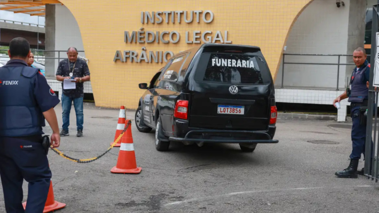 Carro funerário preto com a palavra “FUNERÁRIA” estaciona em frente ao prédio do Instituto Médico Legal Afrânio Peixoto, no Rio de Janeiro. Três homens, incluindo um policial e um agente de segurança, estão próximos ao veículo, enquanto cones laranja delimitam a área de acesso