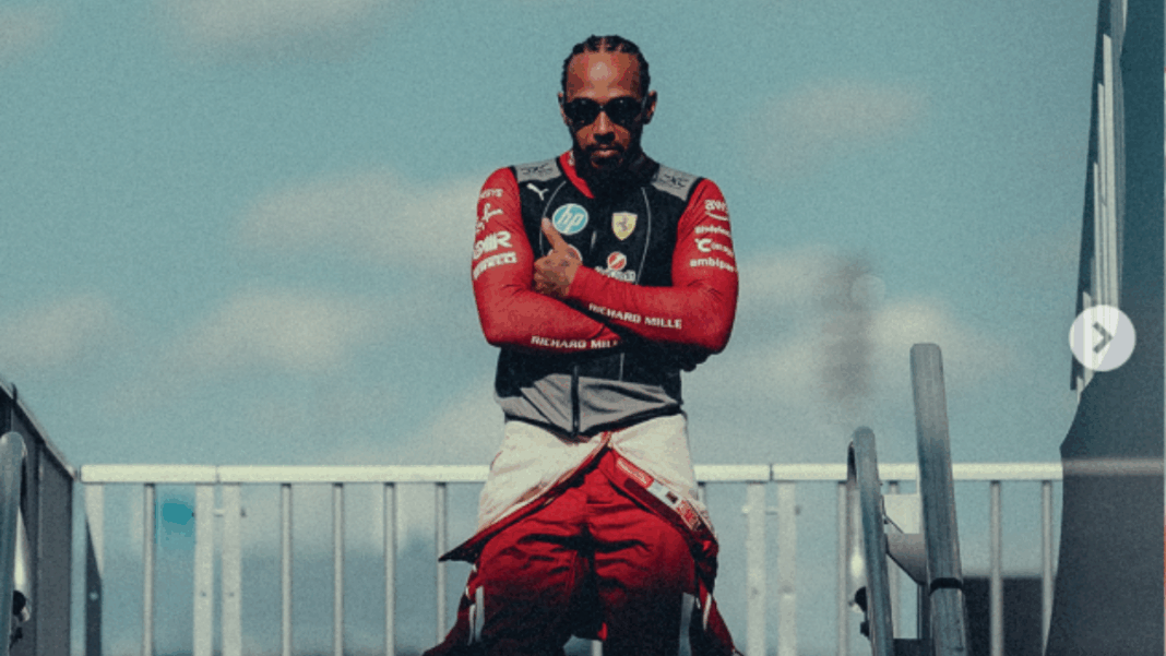 Homem com tranças curtas e óculos escuros posa de braços cruzados, vestindo um macacão de corrida vermelho parcialmente aberto e colete com o logotipo da Ferrari. Ele está em um ambiente externo, sob um céu azul com nuvens suaves, entre estruturas metálicas que lembram o paddock de um autódromo. A iluminação destaca seu semblante confiante e postura firme.