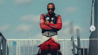 Homem com tranças curtas e óculos escuros posa de braços cruzados, vestindo um macacão de corrida vermelho parcialmente aberto e colete com o logotipo da Ferrari. Ele está em um ambiente externo, sob um céu azul com nuvens suaves, entre estruturas metálicas que lembram o paddock de um autódromo. A iluminação destaca seu semblante confiante e postura firme.