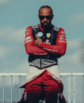 Homem com tranças curtas e óculos escuros posa de braços cruzados, vestindo um macacão de corrida vermelho parcialmente aberto e colete com o logotipo da Ferrari. Ele está em um ambiente externo, sob um céu azul com nuvens suaves, entre estruturas metálicas que lembram o paddock de um autódromo. A iluminação destaca seu semblante confiante e postura firme.