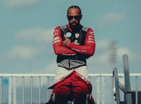 Homem com tranças curtas e óculos escuros posa de braços cruzados, vestindo um macacão de corrida vermelho parcialmente aberto e colete com o logotipo da Ferrari. Ele está em um ambiente externo, sob um céu azul com nuvens suaves, entre estruturas metálicas que lembram o paddock de um autódromo. A iluminação destaca seu semblante confiante e postura firme.