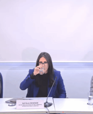 Três pessoas estão sentadas lado a lado em uma mesa de reunião, com microfones e copos de água à frente. À esquerda, um homem de barba e óculos usa terno escuro e camisa branca. No centro, uma mulher de cabelos escuros e lisos, vestindo blazer azul, bebe um copo de água. À direita, outra mulher de óculos, com suéter cinza e camisa branca, fala ao microfone. À frente dela há um notebook com o logotipo “SP Águas” e uma placa de identificação. Ao fundo, uma parede branca com o logotipo do Governo de São Paulo.