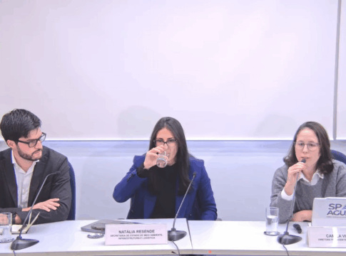 Três pessoas estão sentadas lado a lado em uma mesa de reunião, com microfones e copos de água à frente. À esquerda, um homem de barba e óculos usa terno escuro e camisa branca. No centro, uma mulher de cabelos escuros e lisos, vestindo blazer azul, bebe um copo de água. À direita, outra mulher de óculos, com suéter cinza e camisa branca, fala ao microfone. À frente dela há um notebook com o logotipo “SP Águas” e uma placa de identificação. Ao fundo, uma parede branca com o logotipo do Governo de São Paulo.