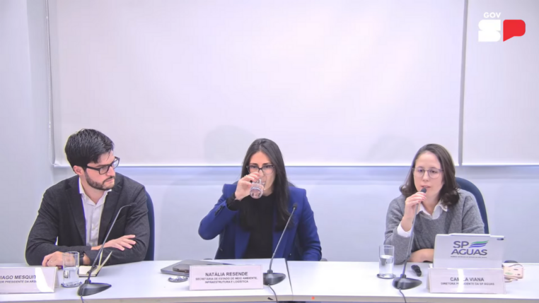 Três pessoas estão sentadas lado a lado em uma mesa de reunião, com microfones e copos de água à frente. À esquerda, um homem de barba e óculos usa terno escuro e camisa branca. No centro, uma mulher de cabelos escuros e lisos, vestindo blazer azul, bebe um copo de água. À direita, outra mulher de óculos, com suéter cinza e camisa branca, fala ao microfone. À frente dela há um notebook com o logotipo “SP Águas” e uma placa de identificação. Ao fundo, uma parede branca com o logotipo do Governo de São Paulo.