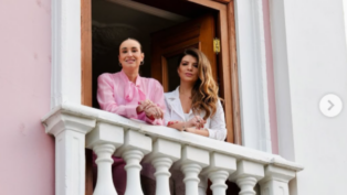 Duas mulheres estão em uma sacada branca de estilo clássico, sorrindo e apoiadas no parapeito. A mulher à esquerda veste uma blusa rosa e tem os cabelos presos, enquanto a da direita usa roupa branca e tem cabelos longos e soltos. Atrás delas há uma porta de madeira aberta e uma parede externa rosa-clara.