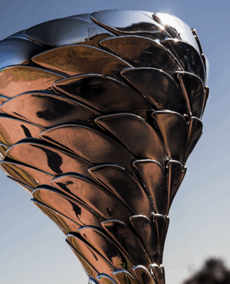 Troféu metálico prateado com design moderno, formado por placas sobrepostas que lembram escamas ou pétalas. Ele está exposto sobre uma base preta, com um autódromo ao fundo sob céu azul e luz solar intensa, sugerindo um evento de corrida ou premiação esportiva.