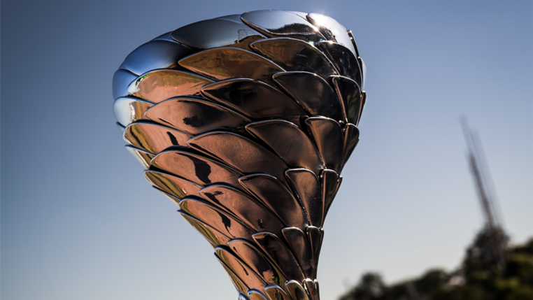 Troféu metálico prateado com design moderno, formado por placas sobrepostas que lembram escamas ou pétalas. Ele está exposto sobre uma base preta, com um autódromo ao fundo sob céu azul e luz solar intensa, sugerindo um evento de corrida ou premiação esportiva.
