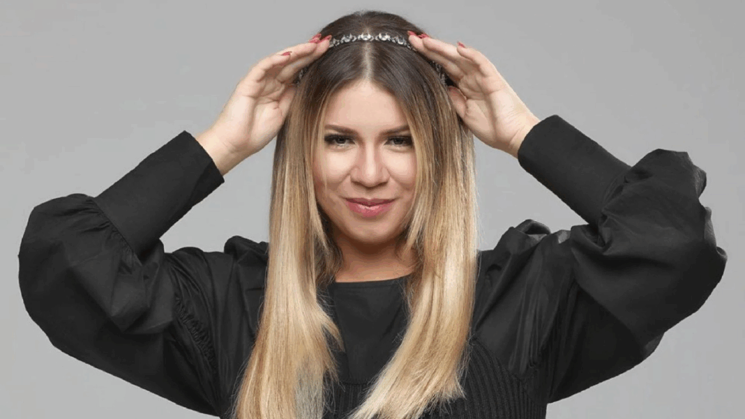 Mulher de cabelos lisos e loiros posa sorrindo levemente diante de um fundo cinza. Ela usa uma blusa preta de mangas bufantes e segura uma tiara prateada sobre a cabeça com as duas mãos.