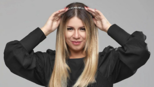 Mulher de cabelos lisos e loiros posa sorrindo levemente diante de um fundo cinza. Ela usa uma blusa preta de mangas bufantes e segura uma tiara prateada sobre a cabeça com as duas mãos.