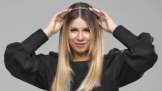 Mulher de cabelos lisos e loiros posa sorrindo levemente diante de um fundo cinza. Ela usa uma blusa preta de mangas bufantes e segura uma tiara prateada sobre a cabeça com as duas mãos.